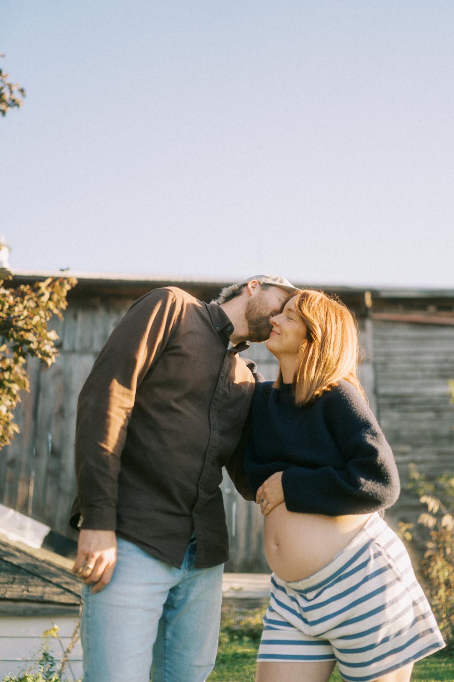 A pregnant couple sharing a tender moment outdoors with the man kissing the woman's forehead, showcasing love and anticipation amidst a rustic background.