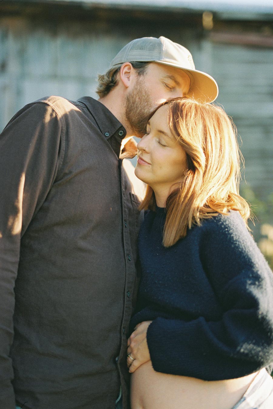 A couple embracing outdoors, with the man kissing the woman's forehead as she cradles her baby bump, capturing a tender moment of love and anticipation during pregnancy.