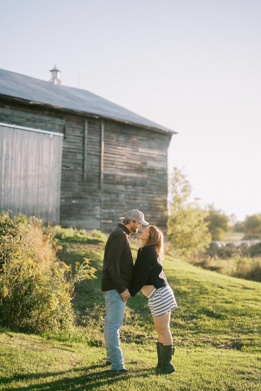 Couple standing affectionately in a grassy field near a rustic wooden barn under a clear sky.