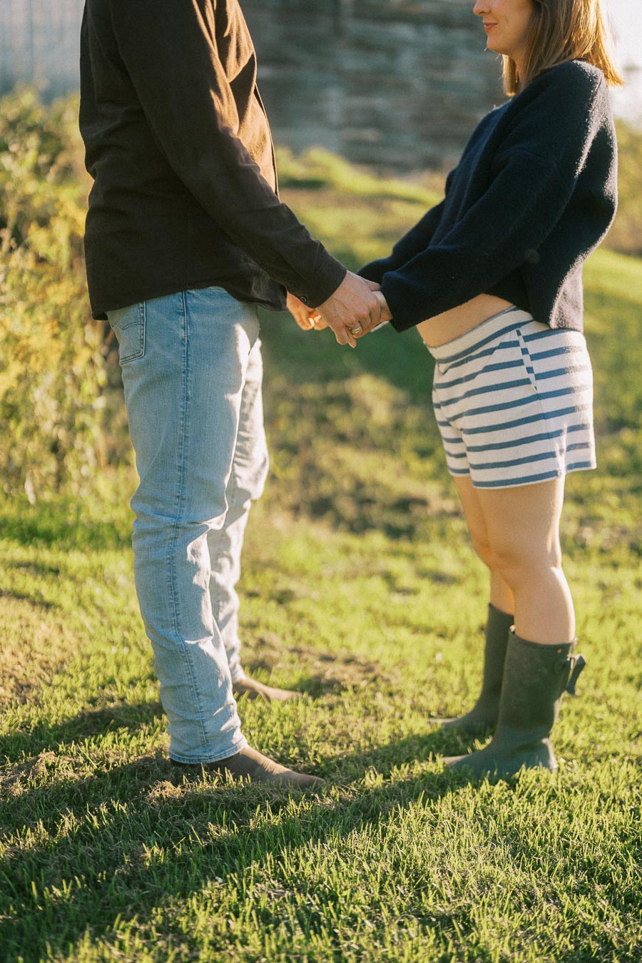 A couple holding hands in a sunny garden, with one person wearing a dark sweater and striped shorts, and the other in jeans and a long-sleeve shirt.