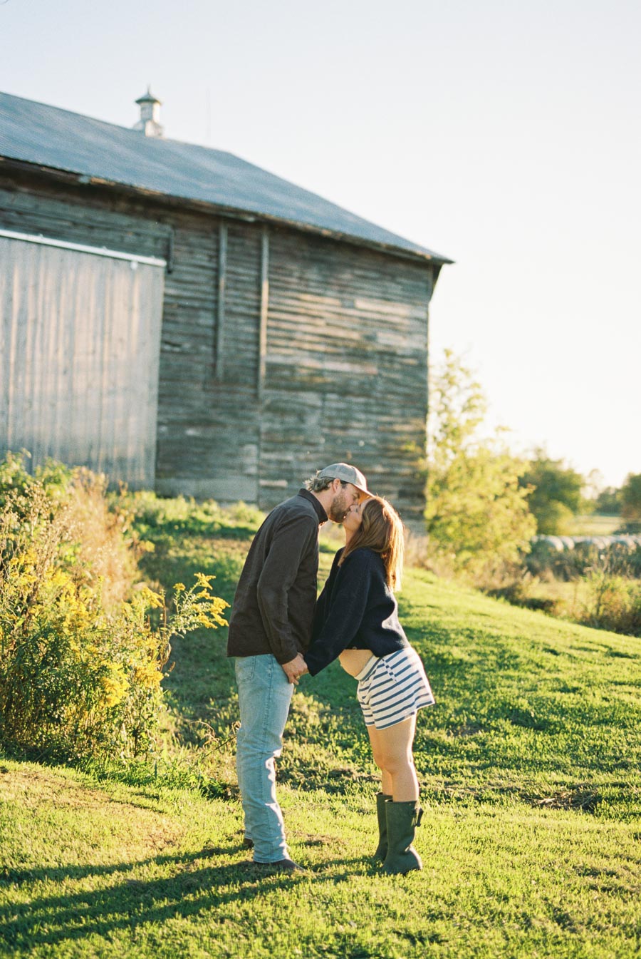 A couple sharing a kiss in a sunny rural setting, standing near a rustic barn with lush greenery and wildflowers, on a bright, clear day.