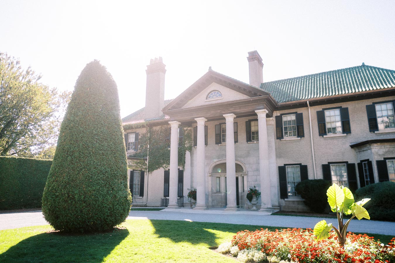 Exterior view of a historic mansion with large white columns, surrounded by well-manicured greenery and colorful flowers under a bright, sunny sky.