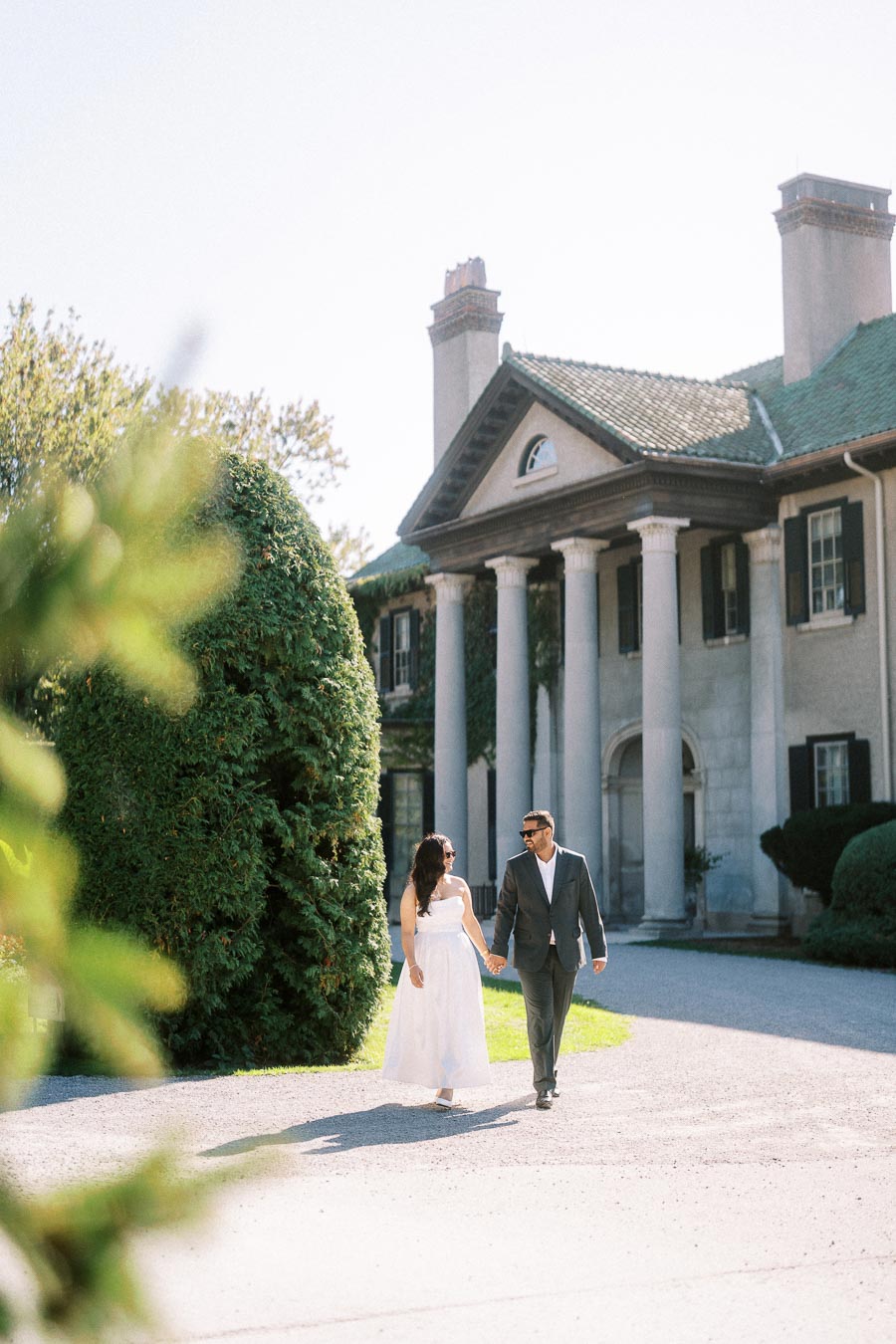 A couple holding hands and walking near a large, elegant manor house with tall white columns and lush greenery.