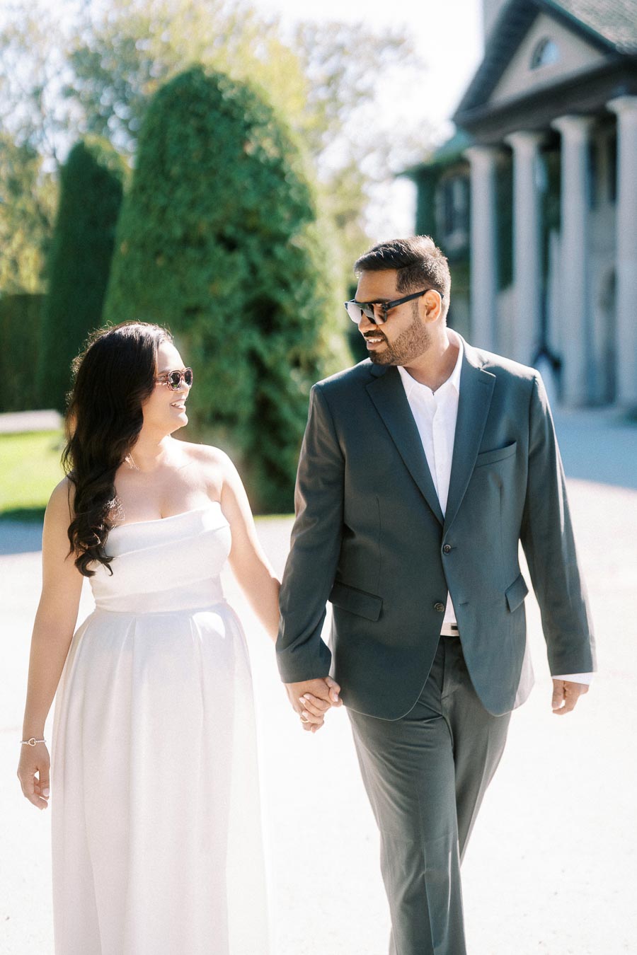 Elegant couple walking hand in hand outdoors, dressed in formal attire with sunglasses, surrounded by greenery and classical architecture, exuding happiness and style.