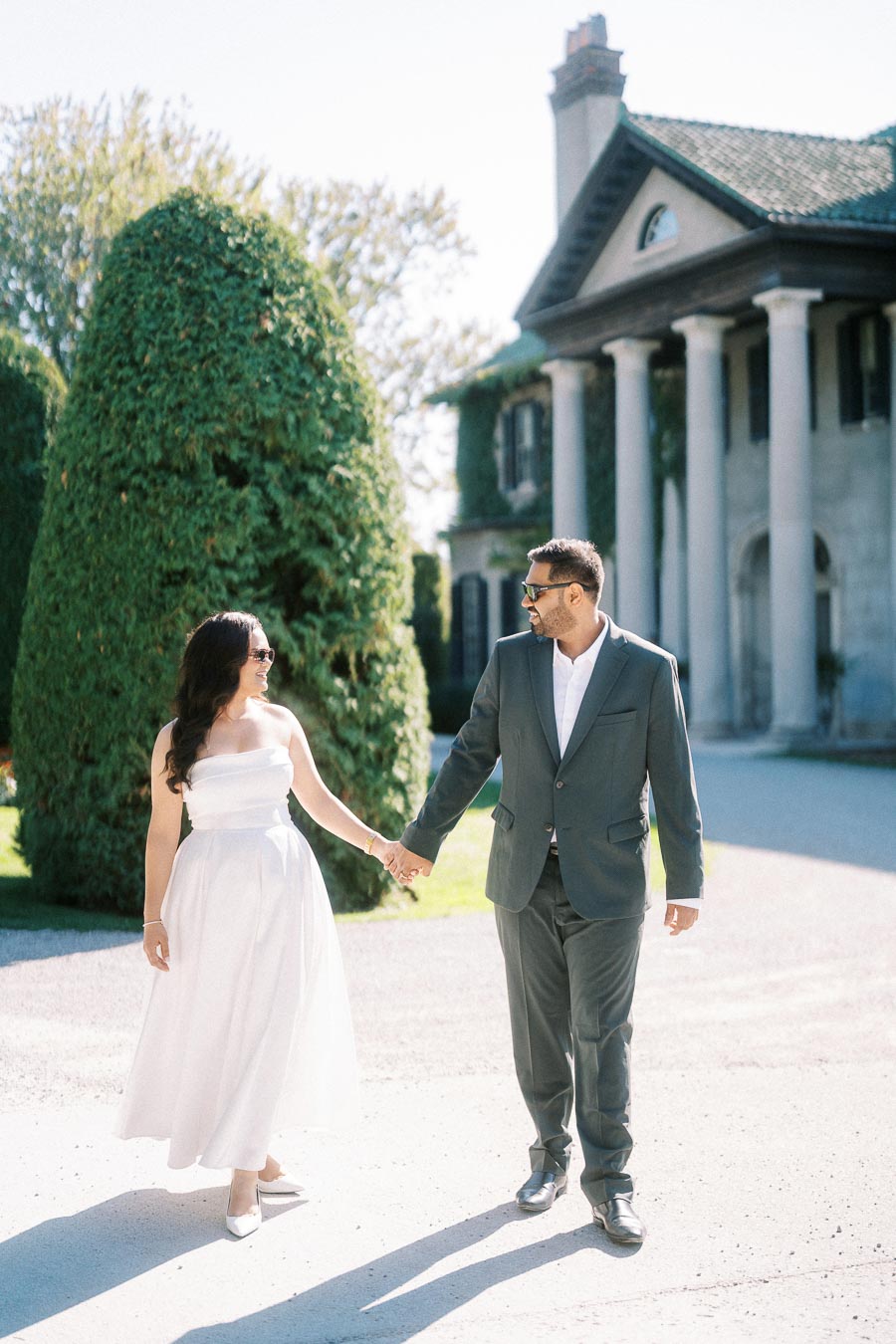 A couple holding hands in front of a stately mansion with tall, manicured trees, both dressed elegantly; the woman in a flowing white dress and the man in a suit, both wearing sunglasses, enjoying a sunny day.