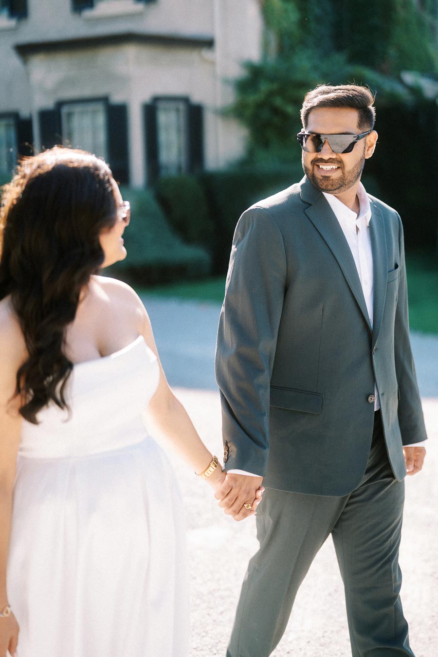 Elegant couple holding hands and smiling, dressed in formal attire with sunglasses, walking outdoors on a sunny day.