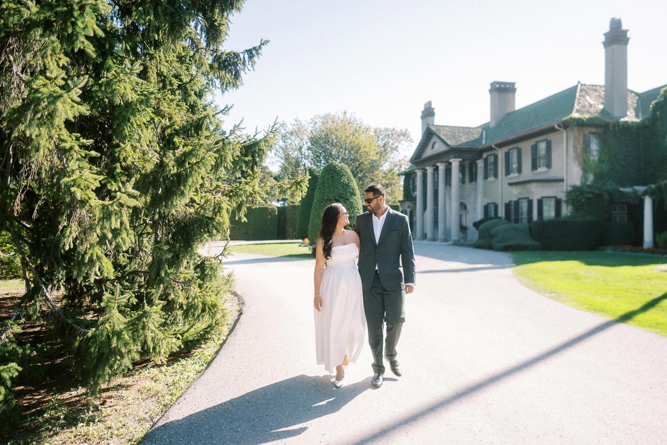 A couple walking hand in hand on a sunny path with lush greenery and a grand mansion in the background, evoking a romantic and elegant atmosphere.