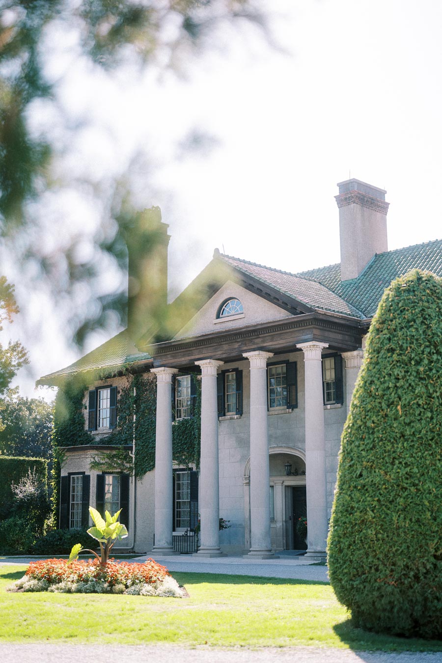 Elegant historic mansion with ivy-covered walls and tall columns, surrounded by lush greenery and vibrant landscaping under a clear sky.