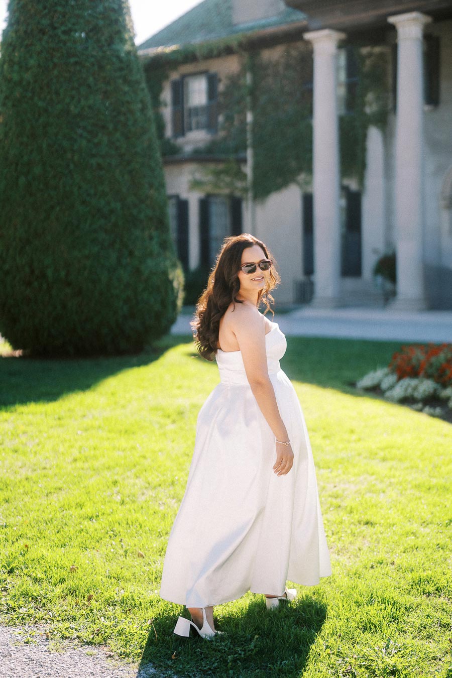 A woman in a white strapless dress and sunglasses stands on a sunlit lawn in front of a classic building with columns and ivy-covered walls.