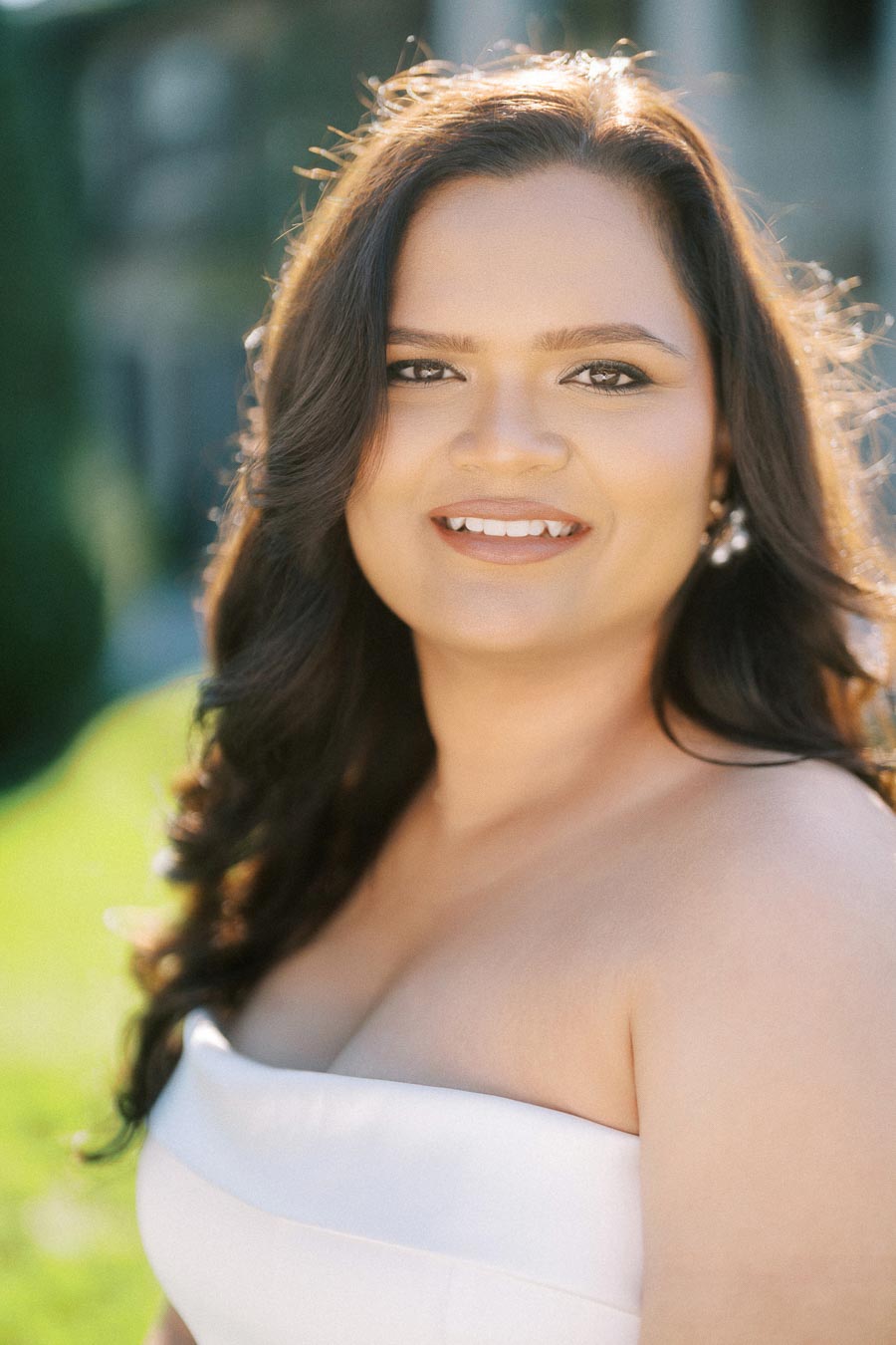Portrait of a woman in a white dress smiling outdoors with soft sunlight highlighting her hair and a blurred green background.