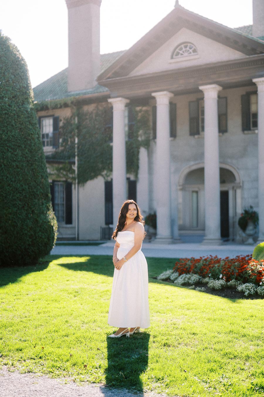 Woman in a white dress standing on a sunlit lawn in front of a historic building with columns and landscaped gardens, conveying elegance and tranquility.