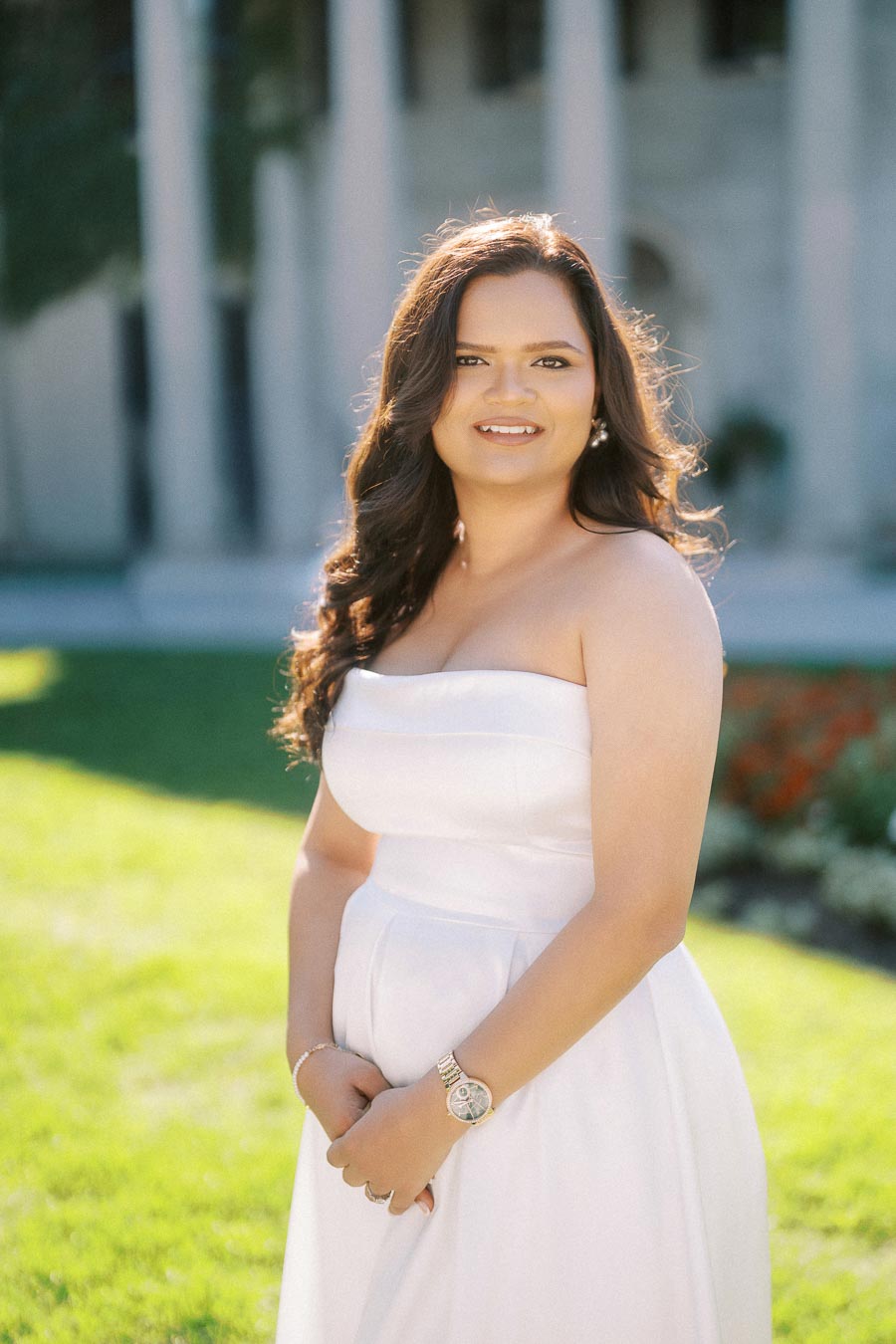 Young woman in an elegant white dress posing outdoors in a garden setting, with a classic building in the background, under bright, sunny weather.