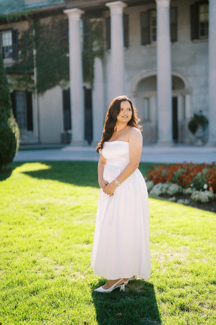 A woman in an elegant white dress stands on a sunlit lawn in front of a stately building with columns and green vines, surrounded by vibrant flowers.