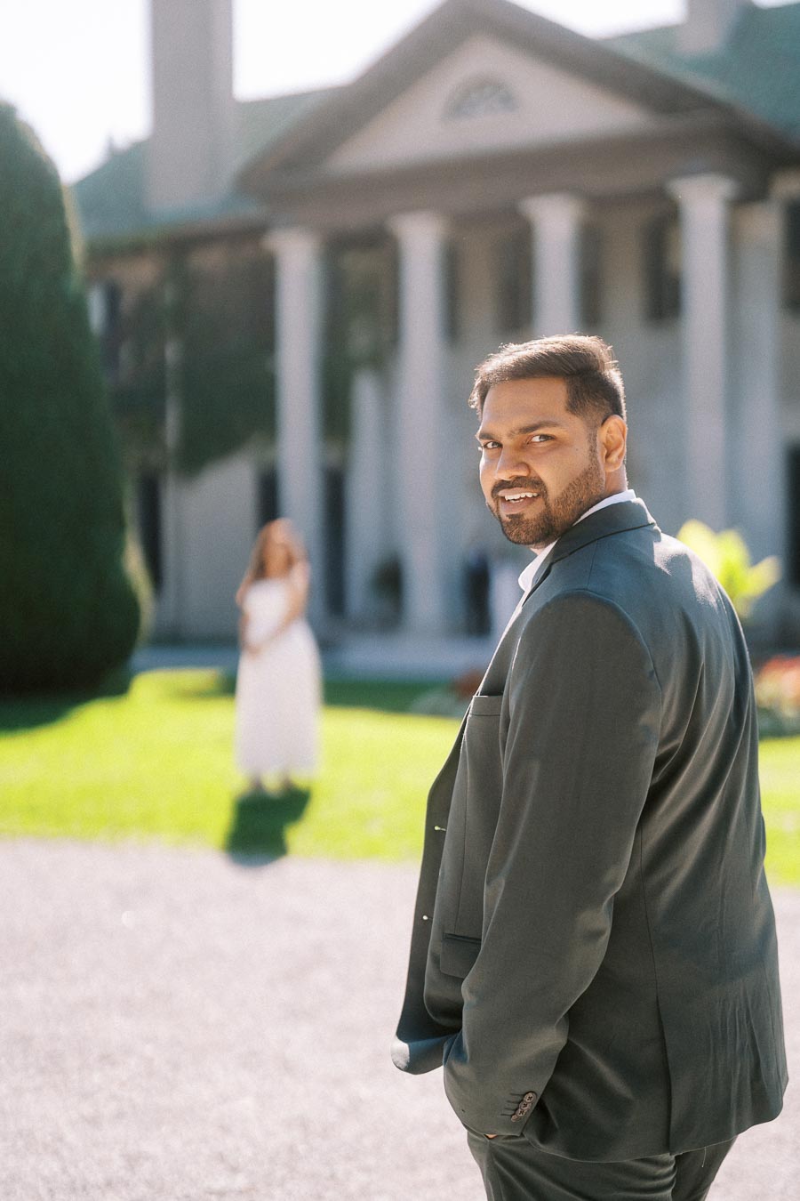 Smiling man in a formal suit outdoors with historic building and blurred figure in the background.