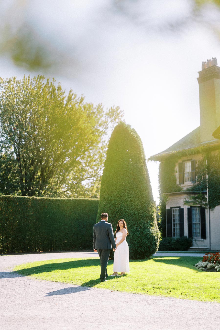 A couple stands in a sunlit garden, with lush greenery and a historic ivy-covered building in the background, creating a romantic atmosphere.