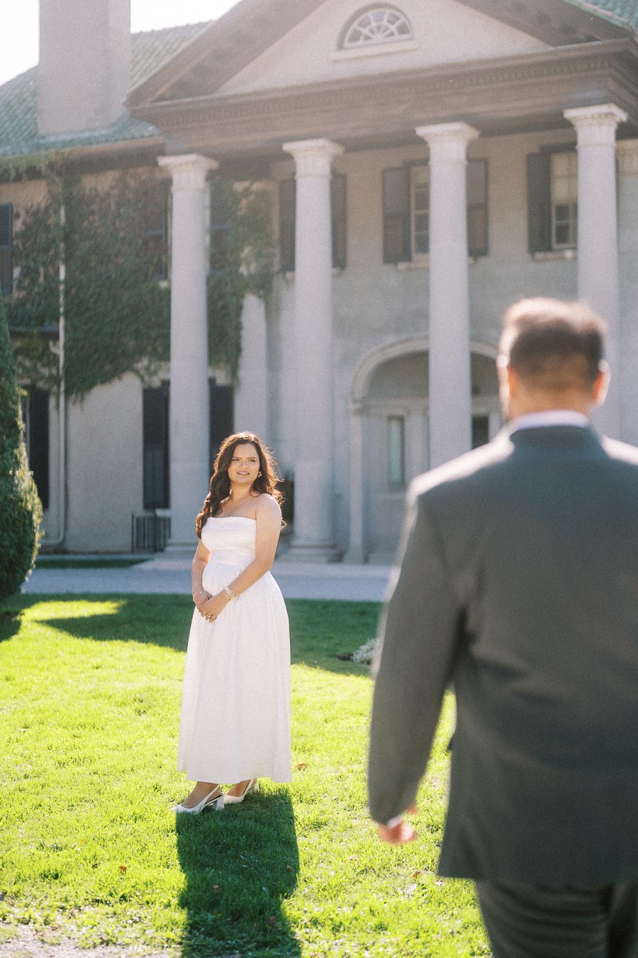 A woman in a white dress stands on a sunlit lawn near a historic building with large columns, facing a man in a suit.