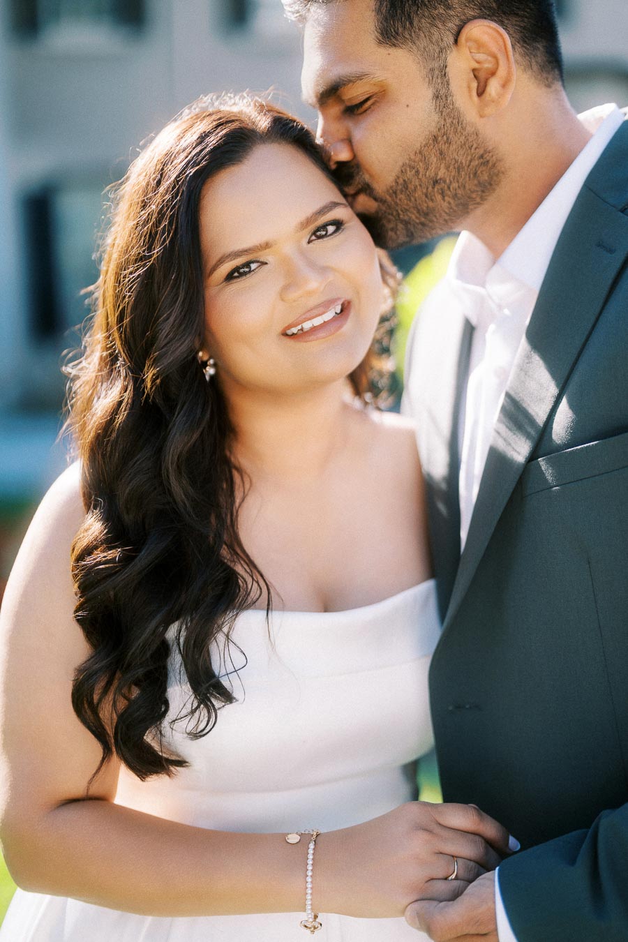 Smiling couple in elegant attire, with the man gently kissing the woman on the forehead during a sunny outdoor engagement photo shoot.