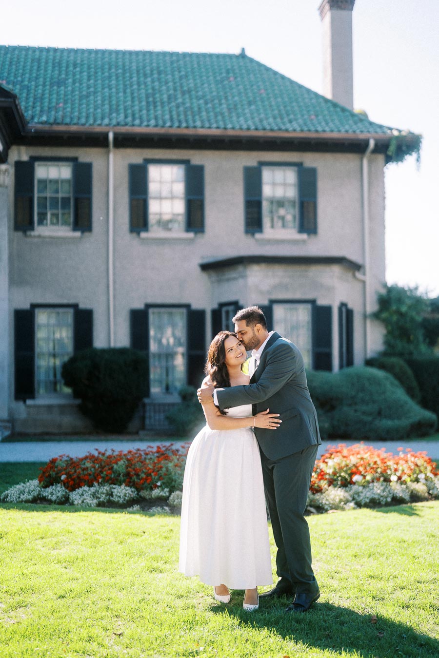 Smiling couple embracing in front of a charming house with a green roof and colorful garden, capturing a joyful and romantic moment