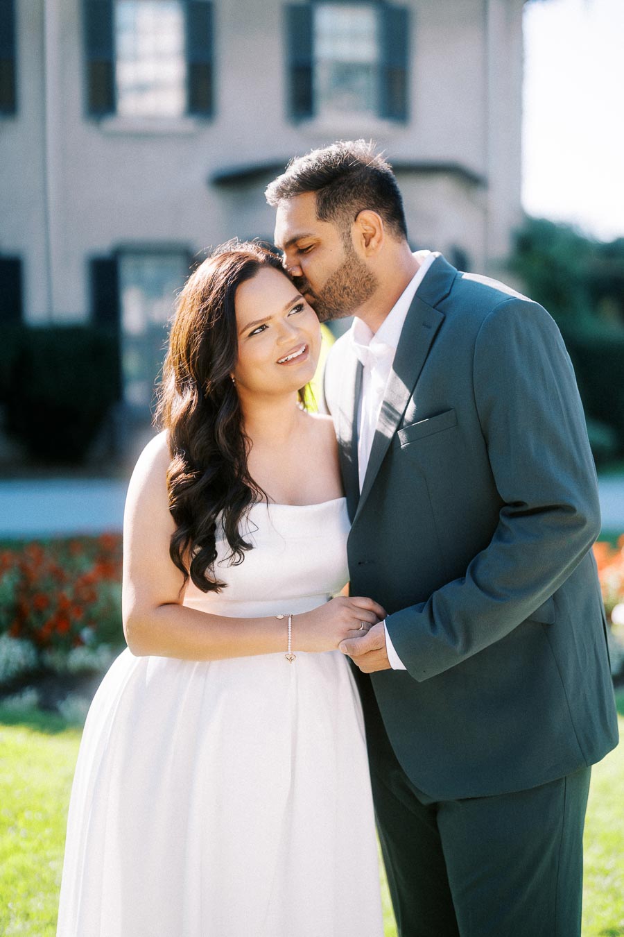 A couple in formal attire shares a tender moment outdoors, with the man kissing the woman's forehead. She is wearing a white dress, and he is in a gray suit. The background features a house and garden on a sunny day.