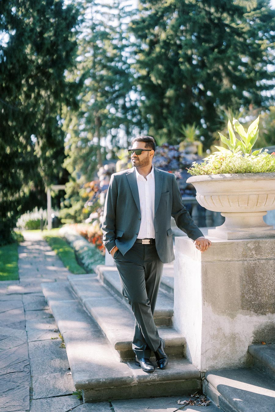 Man in a stylish suit and sunglasses standing outdoors, leaning against a stone railing in a sunny garden setting.