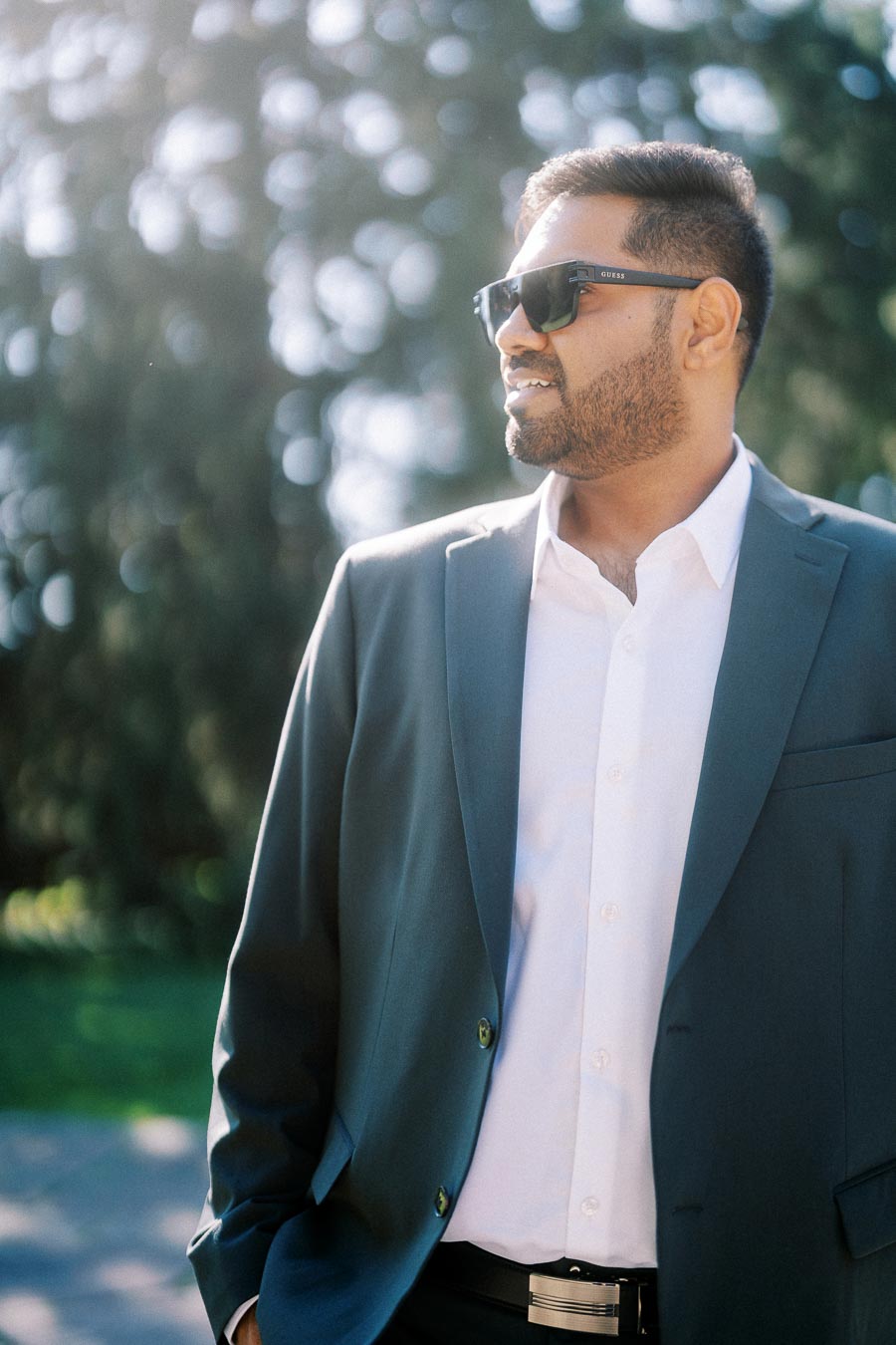 A man in a sharp blue suit and sunglasses standing outdoors on a sunny day, with blurred greenery in the background.