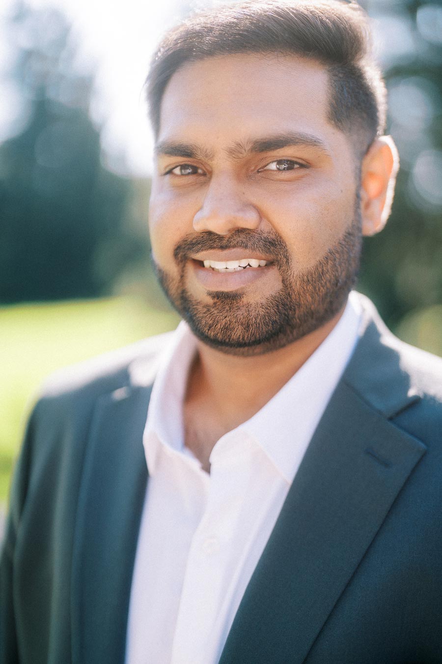 Portrait of a man wearing a dark suit and white shirt, smiling outdoors with a blurred natural background