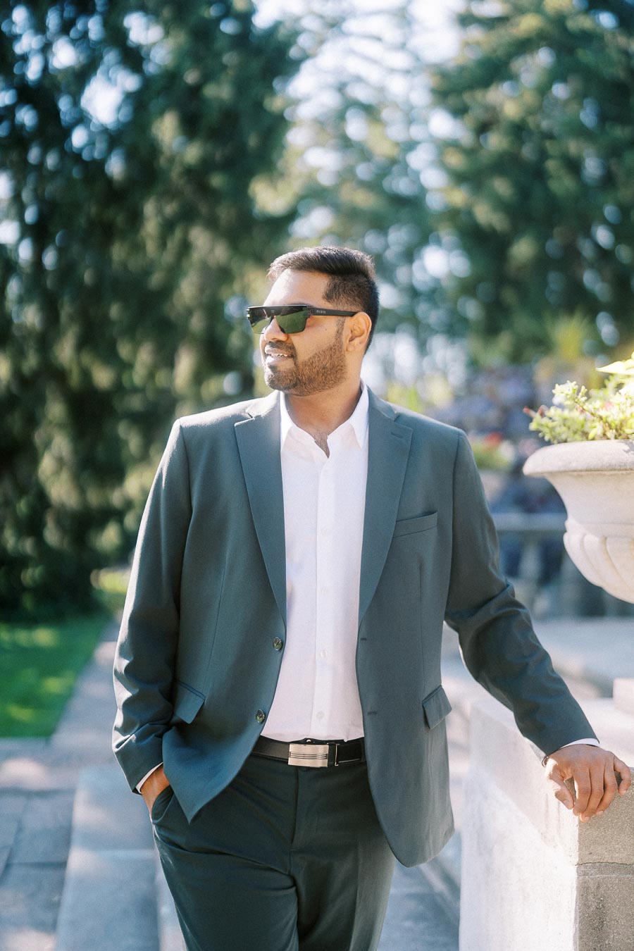 A man in a stylish suit wearing sunglasses stands outdoors in a sunny park, leaning on a stone railing with greenery in the background.