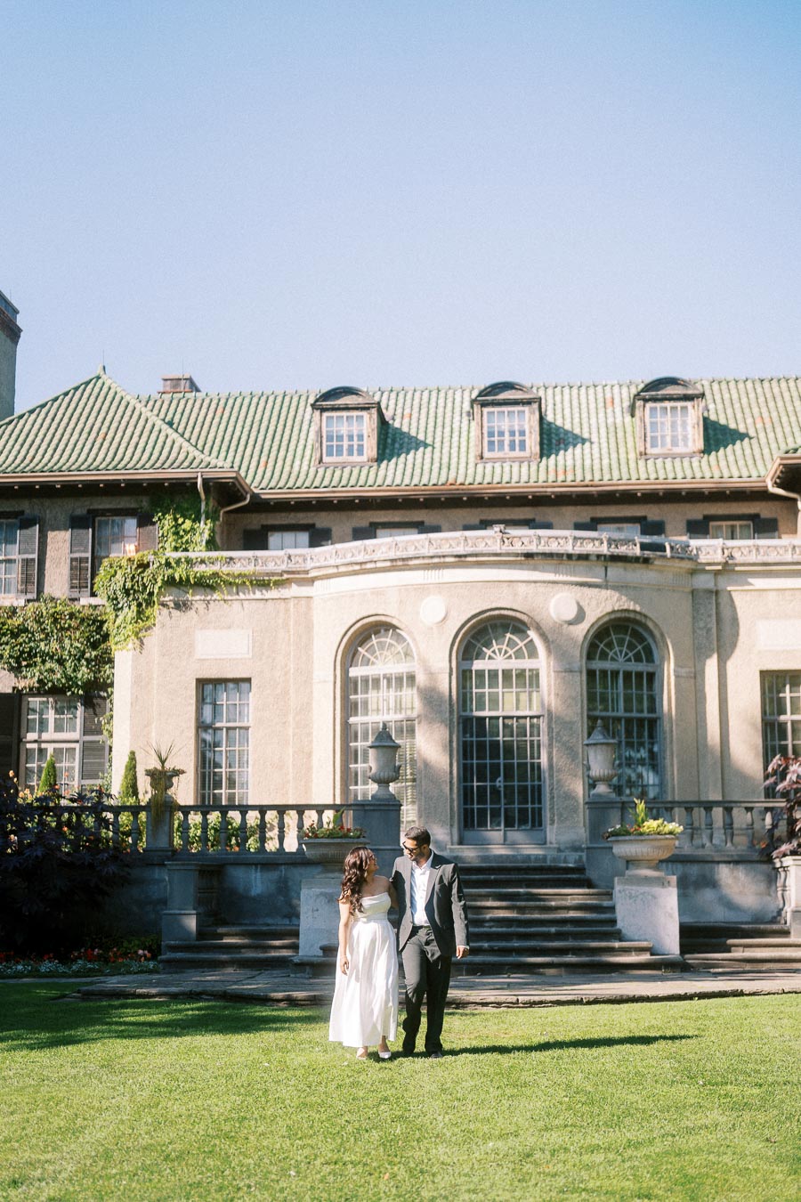 A couple walking on a lush green lawn in front of a grand historic building with large windows and a green tiled roof.