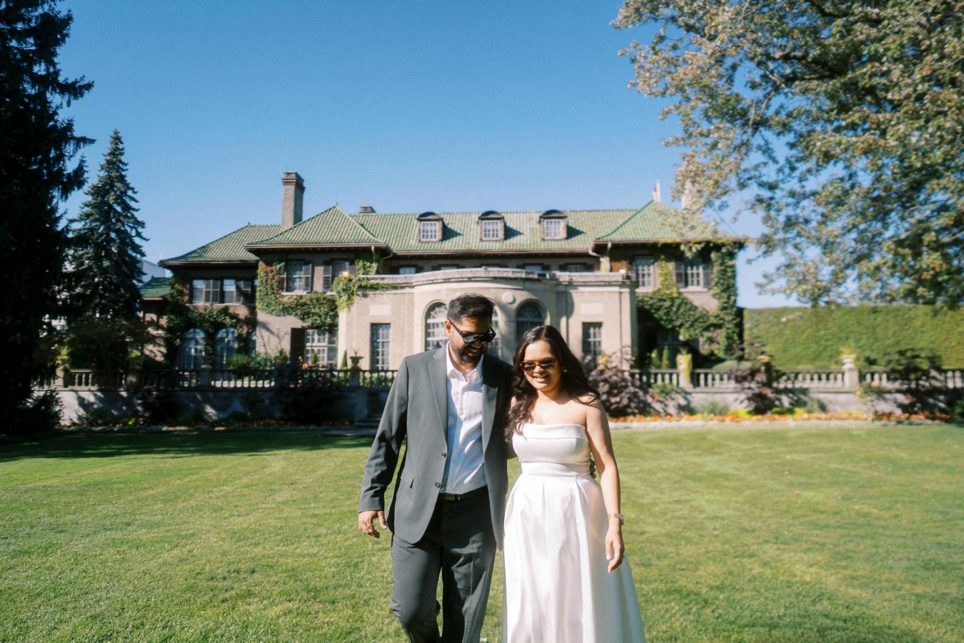 A couple in formal attire walking on a lush green lawn in front of a grand historic mansion on a sunny day.