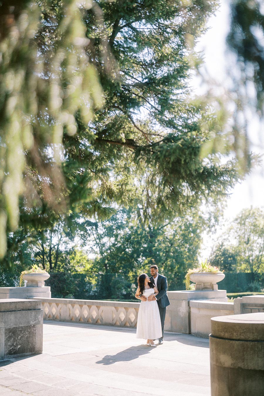 Romantic couple embracing on a scenic outdoor terrace surrounded by lush greenery and elegant stone architecture under a bright, sunlit sky