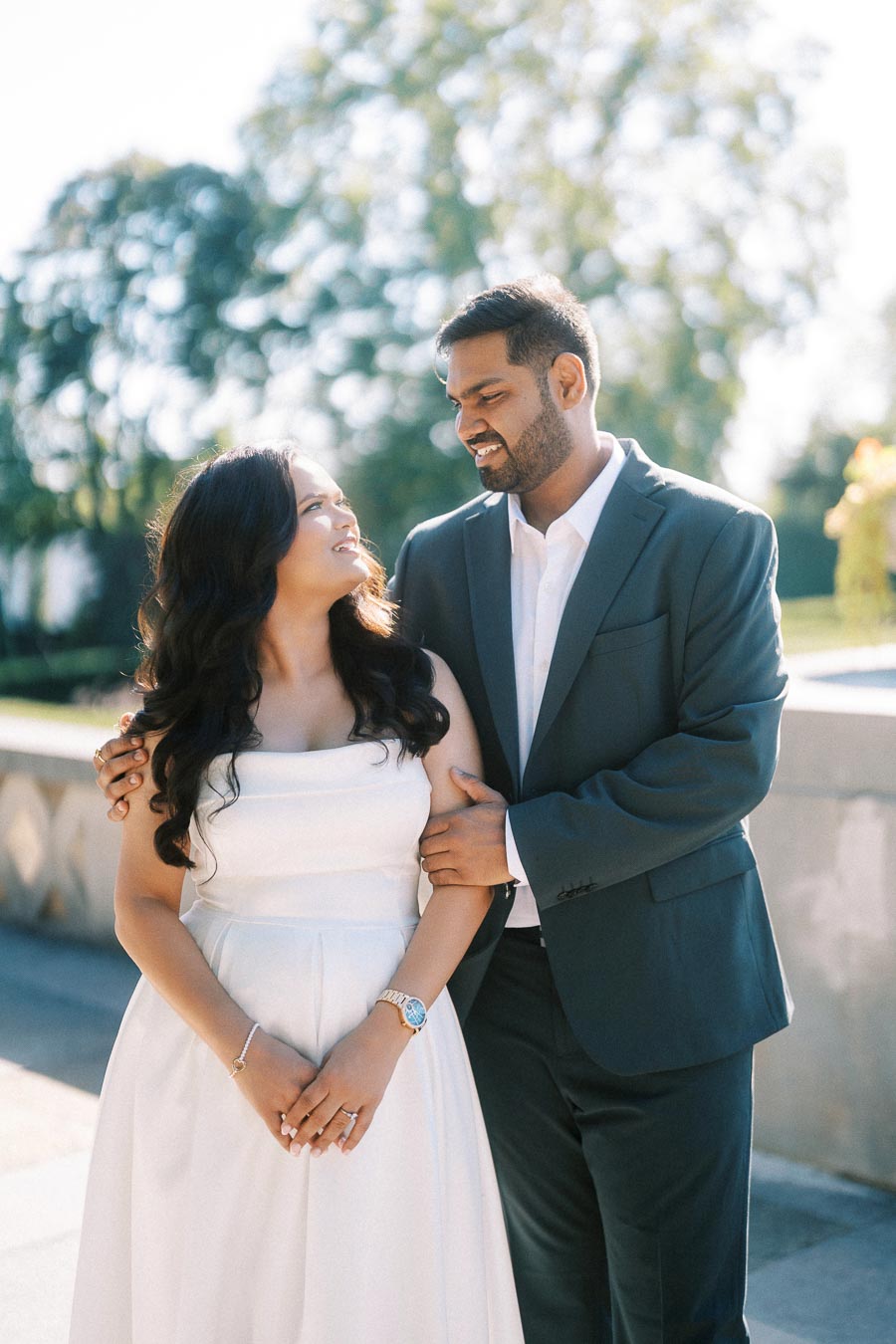 A couple smiling at each other on a sunny day, with the man wearing a dark suit and the woman in a white dress, standing in an outdoor setting with trees in the background.