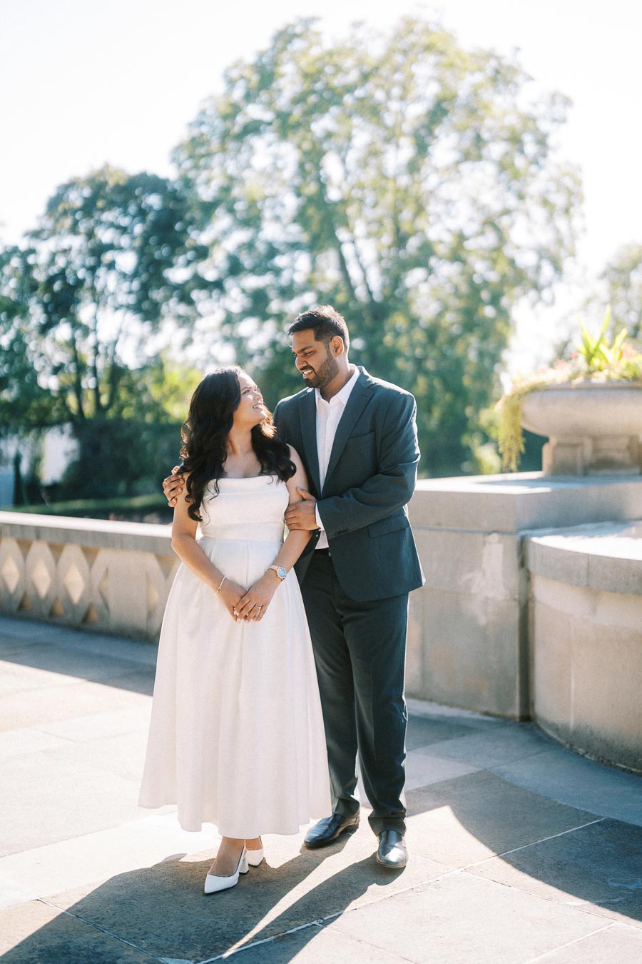A couple smiling at each other, standing in a sunlit outdoor setting with lush greenery in the background. The woman is wearing a white dress, and the man is dressed in a dark suit, creating a romantic and elegant atmosphere.