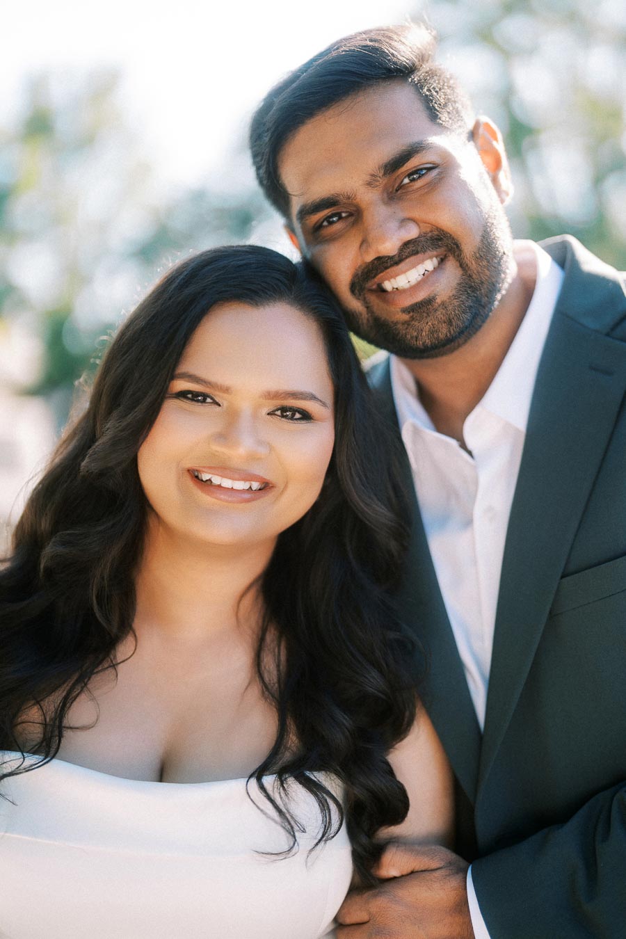 Smiling couple embracing outdoors under sunlight, dressed in formal attire with a blurred natural background.