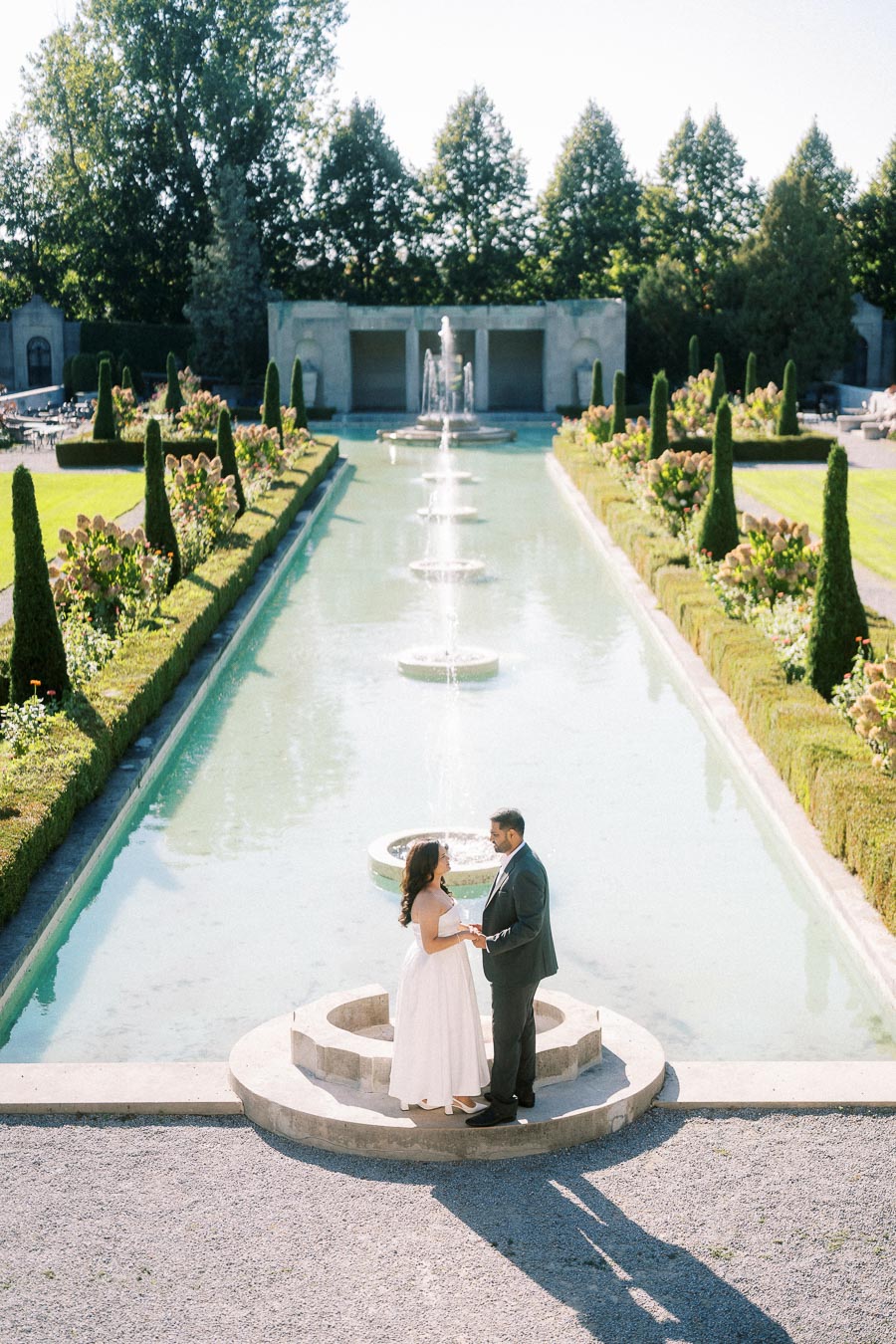 Elegant couple standing by a scenic fountain in a beautifully manicured garden, surrounded by lush greenery and topiary under a clear blue sky.
