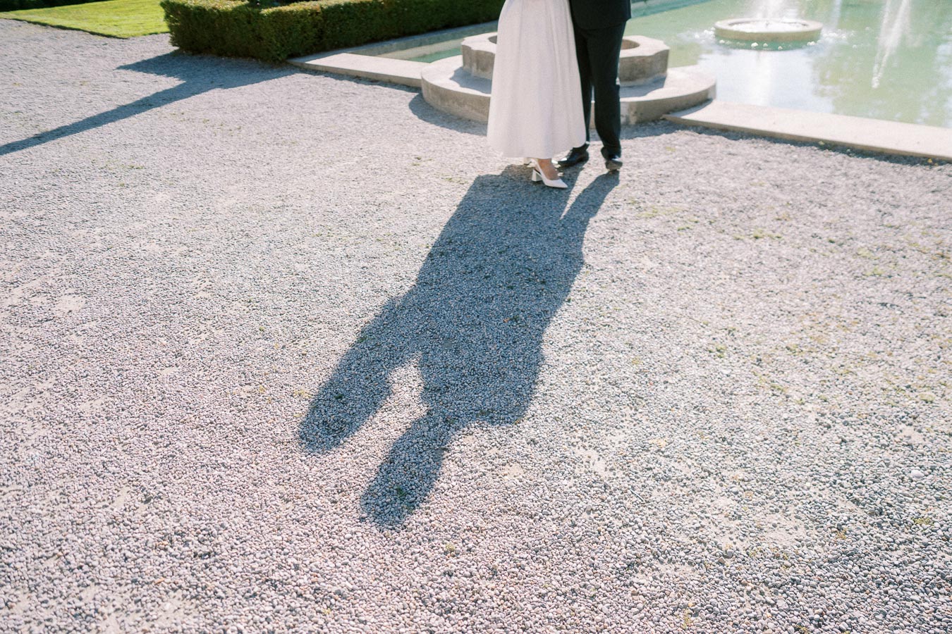 A romantic couple's shadow on gravel near a fountain, capturing a serene moment in an elegant garden setting.