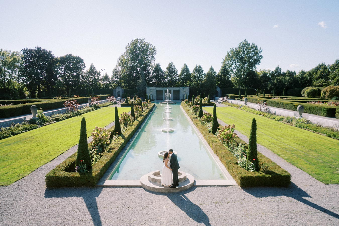 Romantic couple embracing at the edge of a long, elegant fountain in a lush, manicured garden under a clear blue sky, symbolizing love and tranquility.