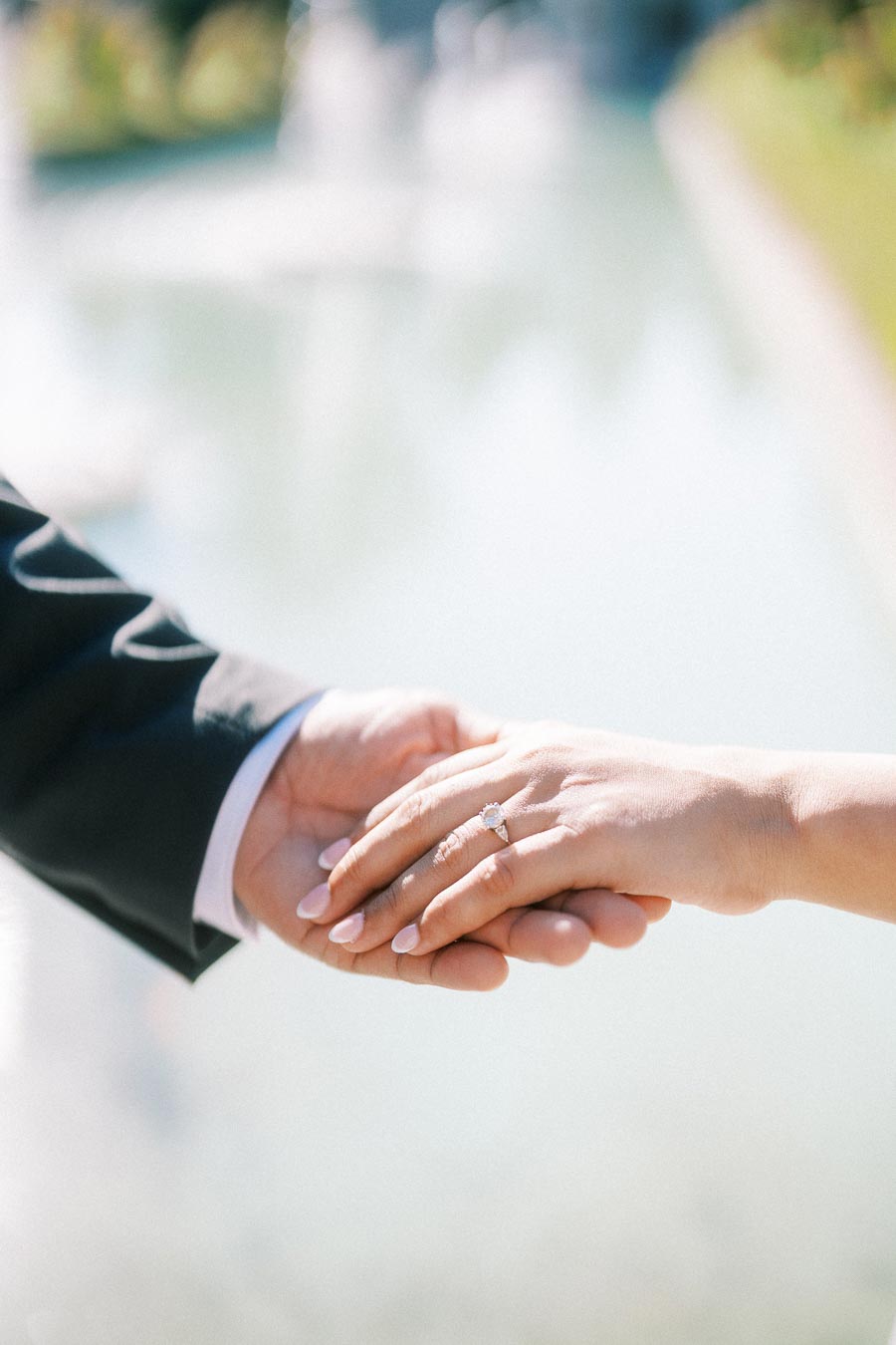 A close-up of a couple holding hands, with an engagement ring visible on the woman's finger, set against a soft, out-of-focus natural background.