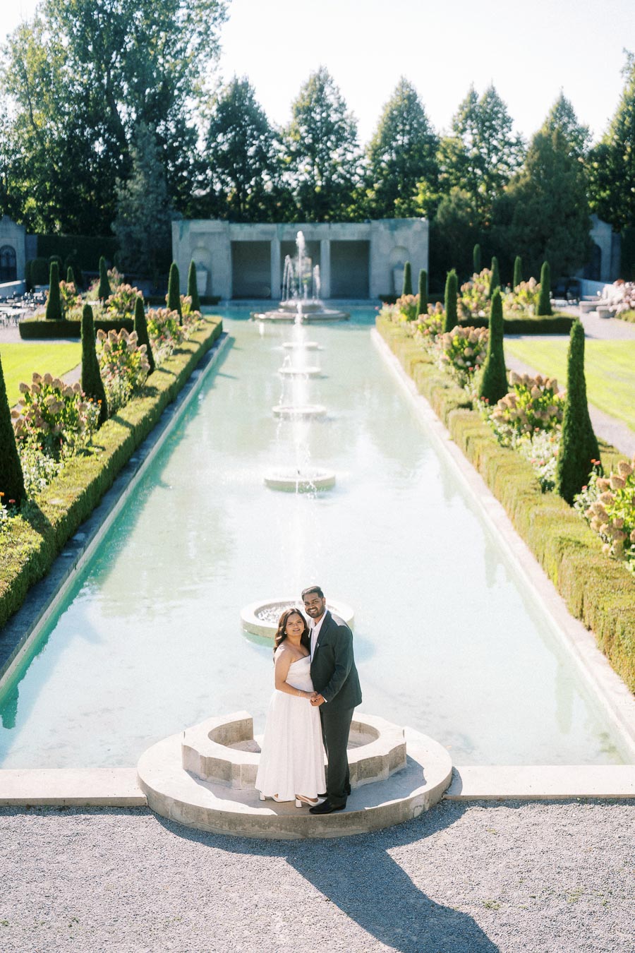 A couple in formal attire poses by a picturesque garden fountain at a historic estate, surrounded by manicured greenery and blooming flowers under a clear blue sky.