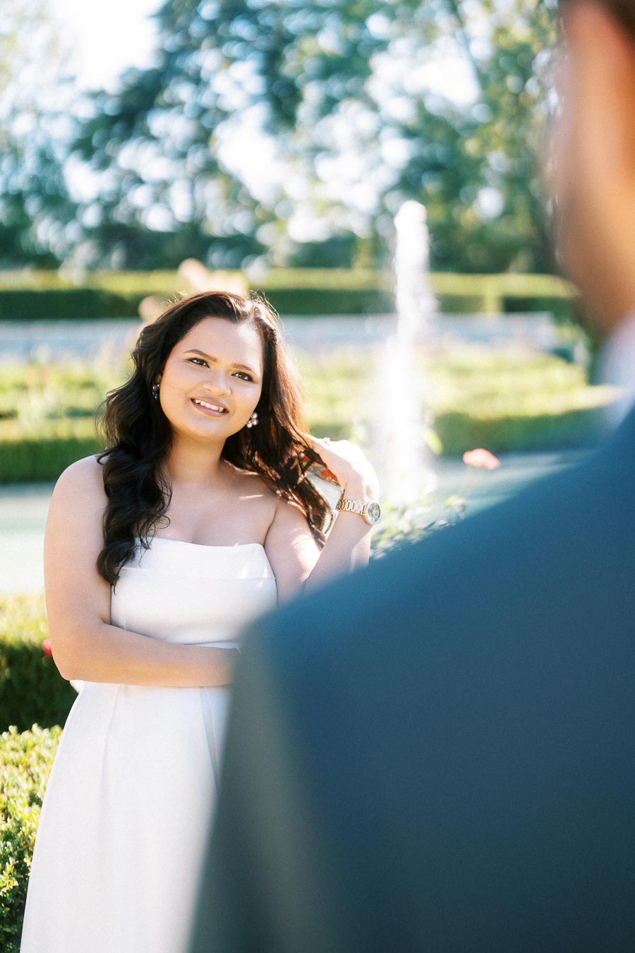 A woman in a white dress smiles in a garden setting with a fountain in the background, partially obscured by the back of another person in a suit.