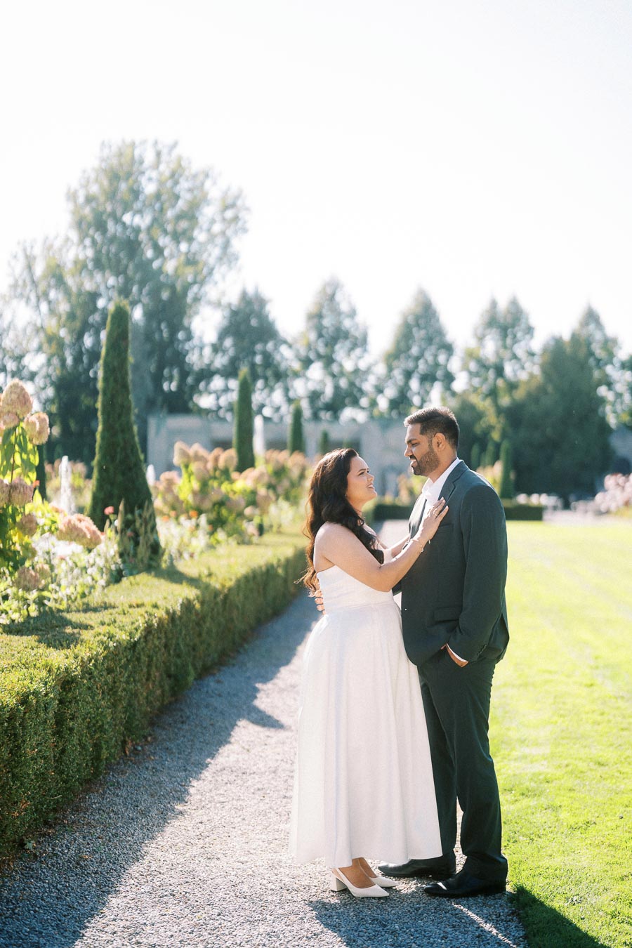 Romantic couple posing in elegant formal wear on a sunny day in a lush garden setting with blooming flowers and trimmed hedges