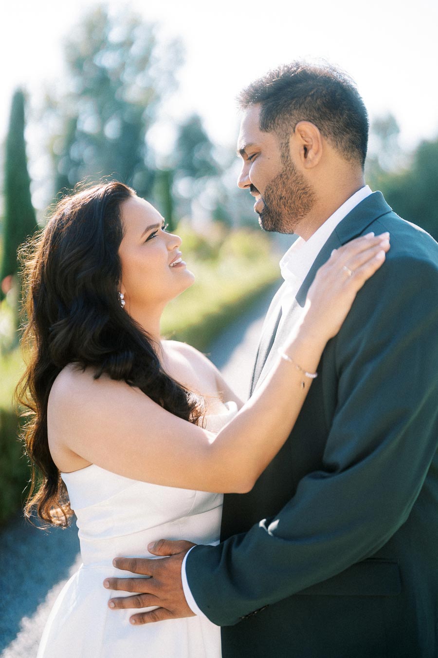Happy couple embracing outdoors with greenery in the background, woman in white dress and man in dark suit smiling at each other, sunny day.