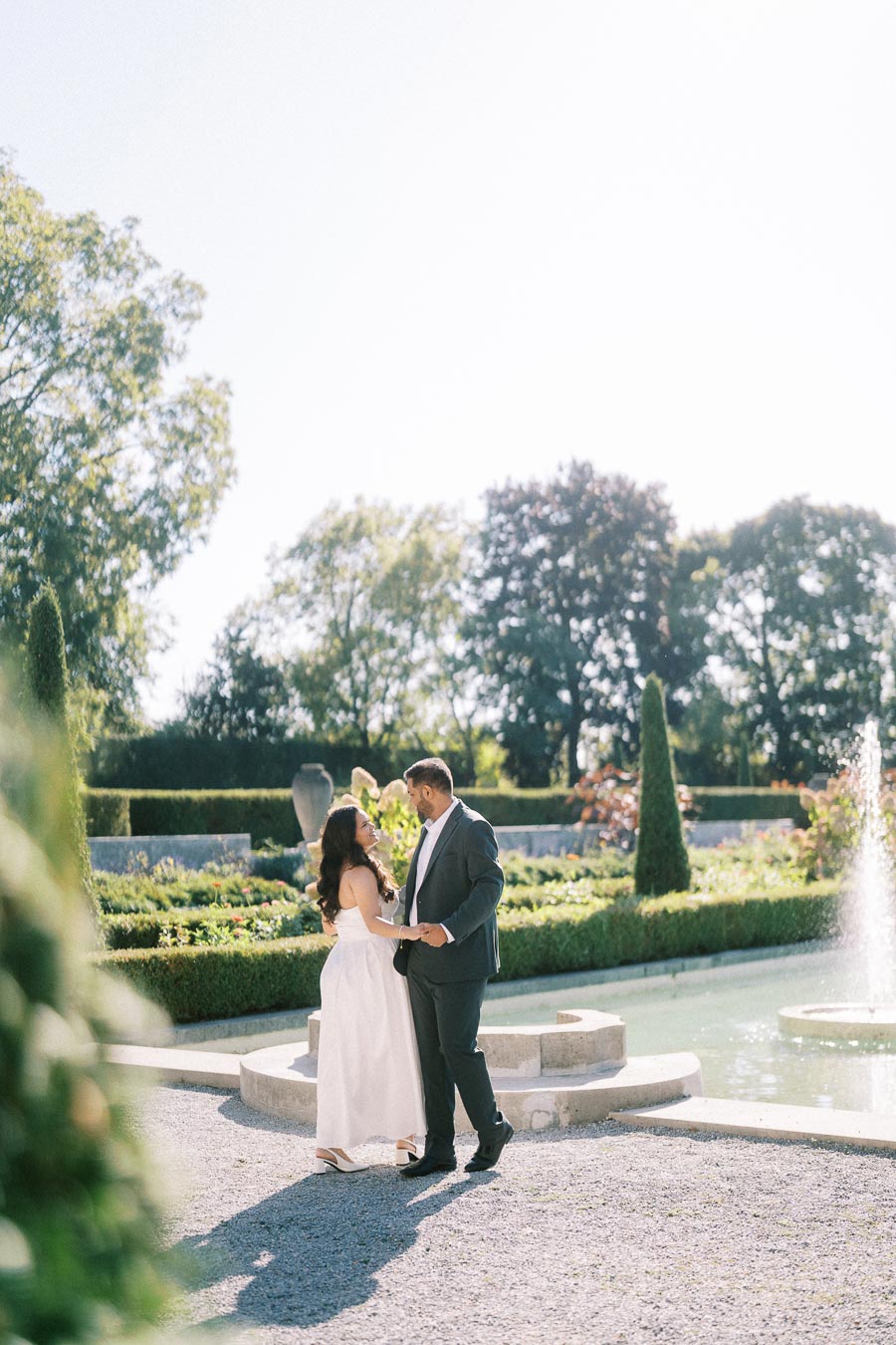 A couple dancing near a fountain in a beautifully landscaped garden under a clear blue sky.