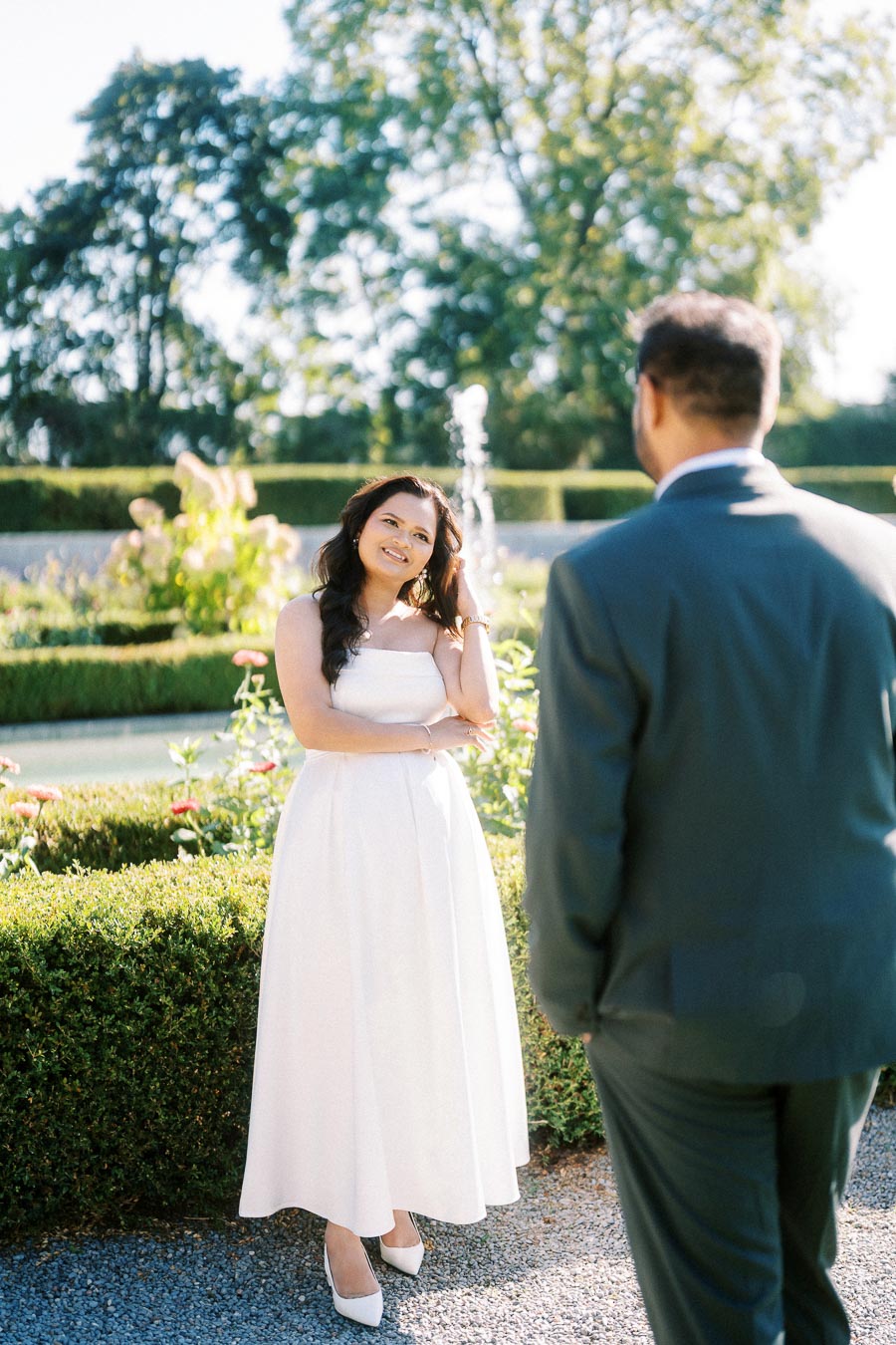 A woman in a white dress standing in a garden, smiling at a man in a suit. The scene includes lush greenery and a fountain in the background, suggesting a romantic outdoor setting.