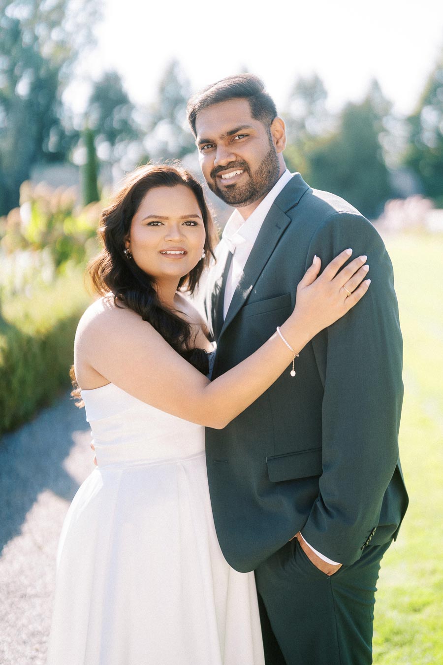 A happy couple embracing outdoors in formal attire; woman in a white dress and man in a suit, with a bright, natural background.