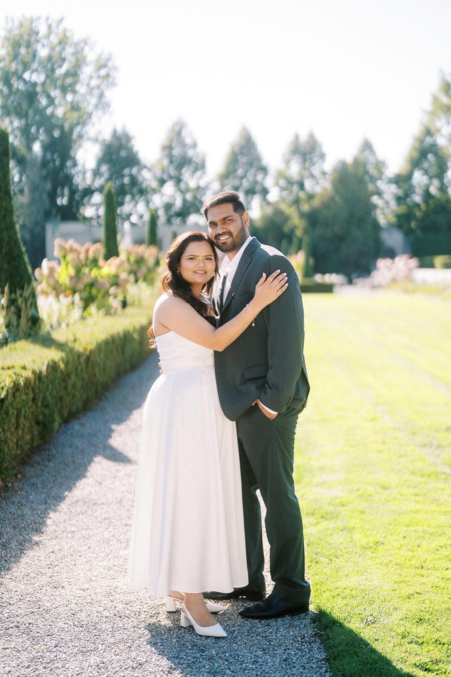 A couple embracing in a sunlit garden, the woman wearing a white dress and the man in a dark suit, surrounded by lush greenery and a beautifully manicured lawn.