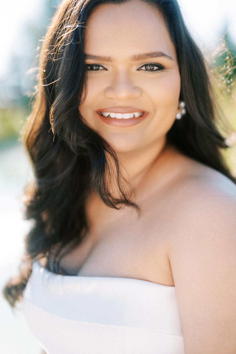 A smiling woman with long dark hair wearing a strapless white top, outdoors in natural sunlight.
