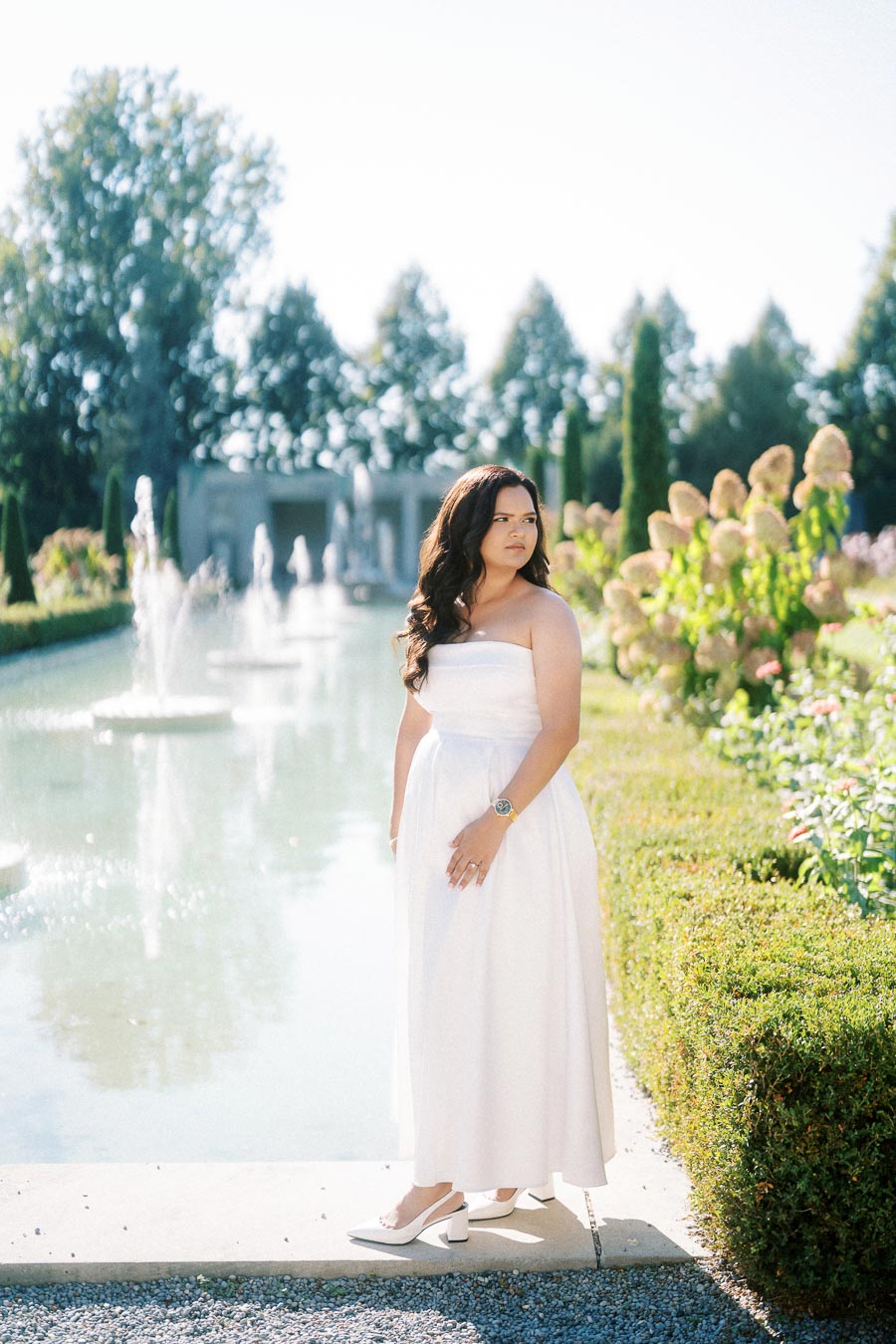 A woman in a white strapless dress stands elegantly beside a reflective pond in a beautifully landscaped garden, with fountains and blooming flowers on a sunny day.