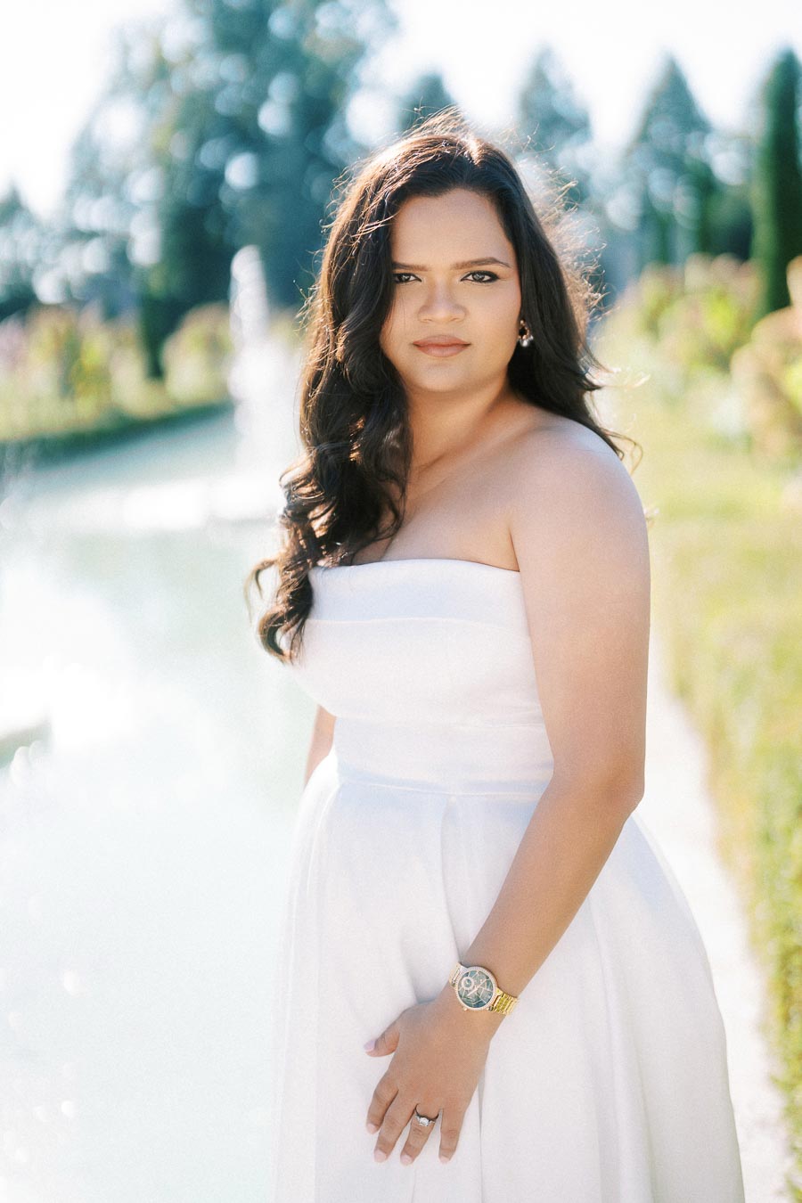 Brunette woman in a white strapless dress standing outdoors by a fountain, surrounded by greenery.