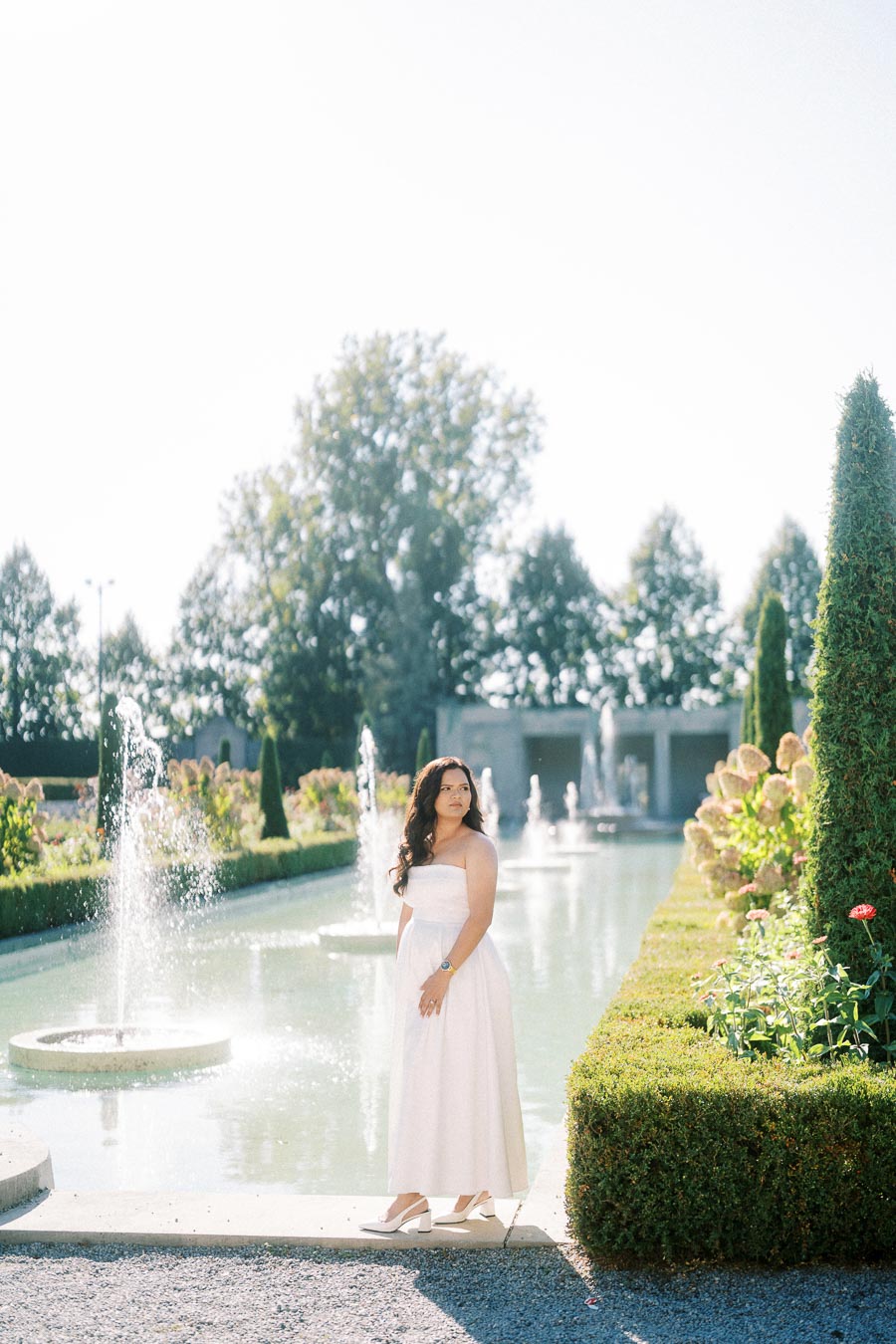 Woman in a white dress standing by a fountain in a sunlit garden with manicured hedges and blooming flowers.
