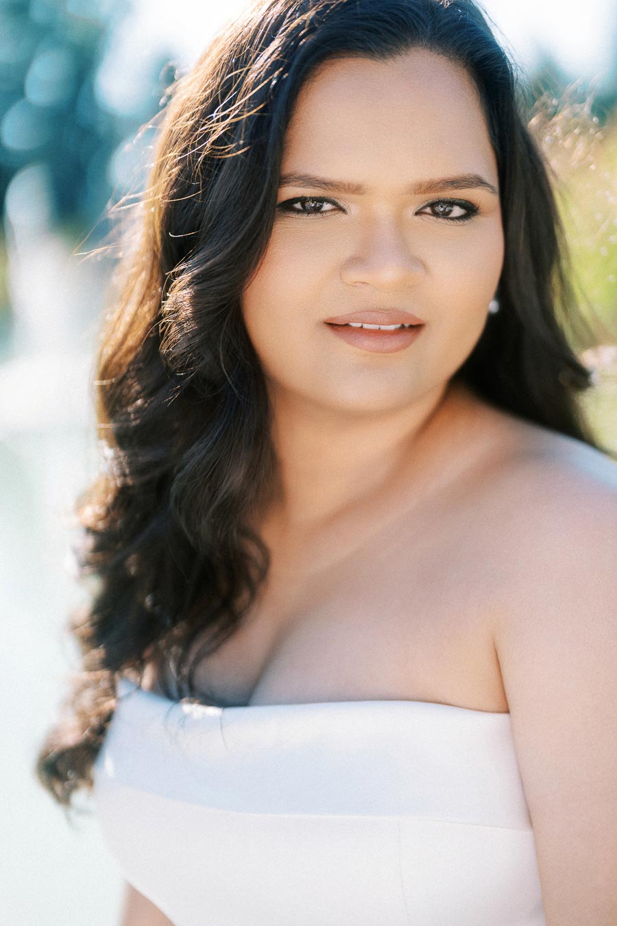 Portrait of a woman with long dark hair and a strapless white dress, smiling softly with a blurred outdoor background.